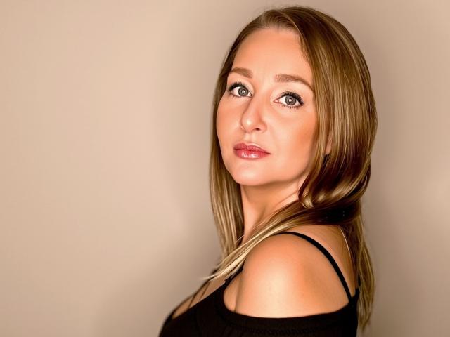 SensualLEA mit blonden Haaren, grünen Augen und pinkem Lippenstift, trägt ein schwarzes Oberteil, blickt über ihre Schulter, sanftes Lächeln, glatte Haut, leicht geöffneter Mund, sexy, attraktiv..