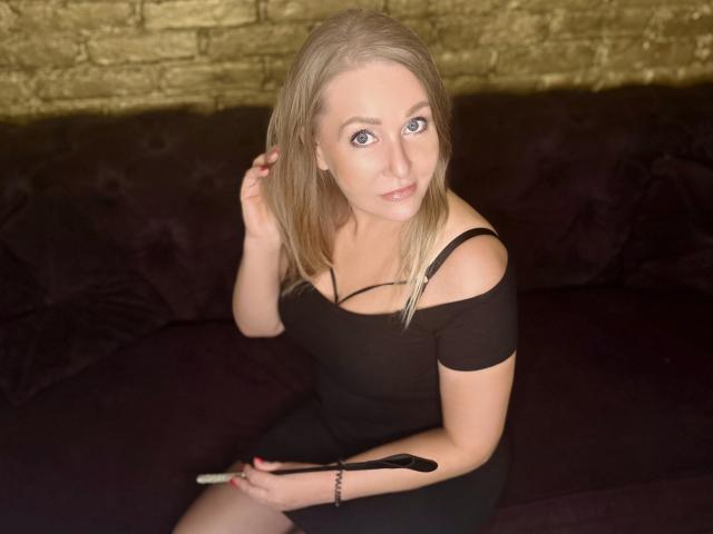 SensualLEA with blonde hair, off-shoulder black dress, holding a whip, sitting on dark purple couch, looking up with blue eyes, slight smile, bracelet on right wrist.
