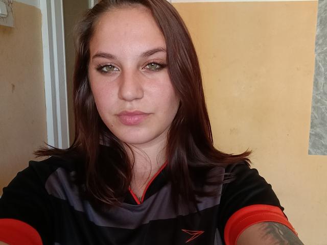 WinnieFalso mit braunen Haaren, grünen Augen und rosafarbenen Lippen, trägt ein schwarzes und rotes Sportshirt und blickt leicht schmollend in die Kamera. Ihr Tattoo ist auf ihrem Arm sichtbar..