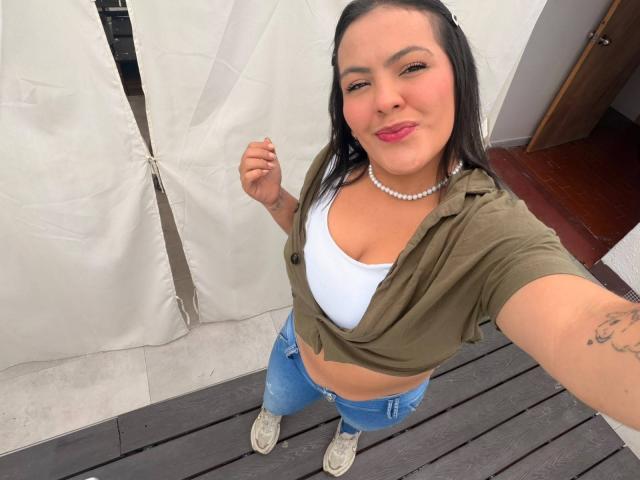 Antonela Vega hat Brüste, die in einem weißen BH zu sehen sind, trägt ein grünes, geknotetes Hemd, blaue Jeans, weiße Turnschuhe, eine Perlenkette und hat dunkles Haar. Sie lächelt mit rotem Lippenstift und steht auf einem hölzernen Deck..