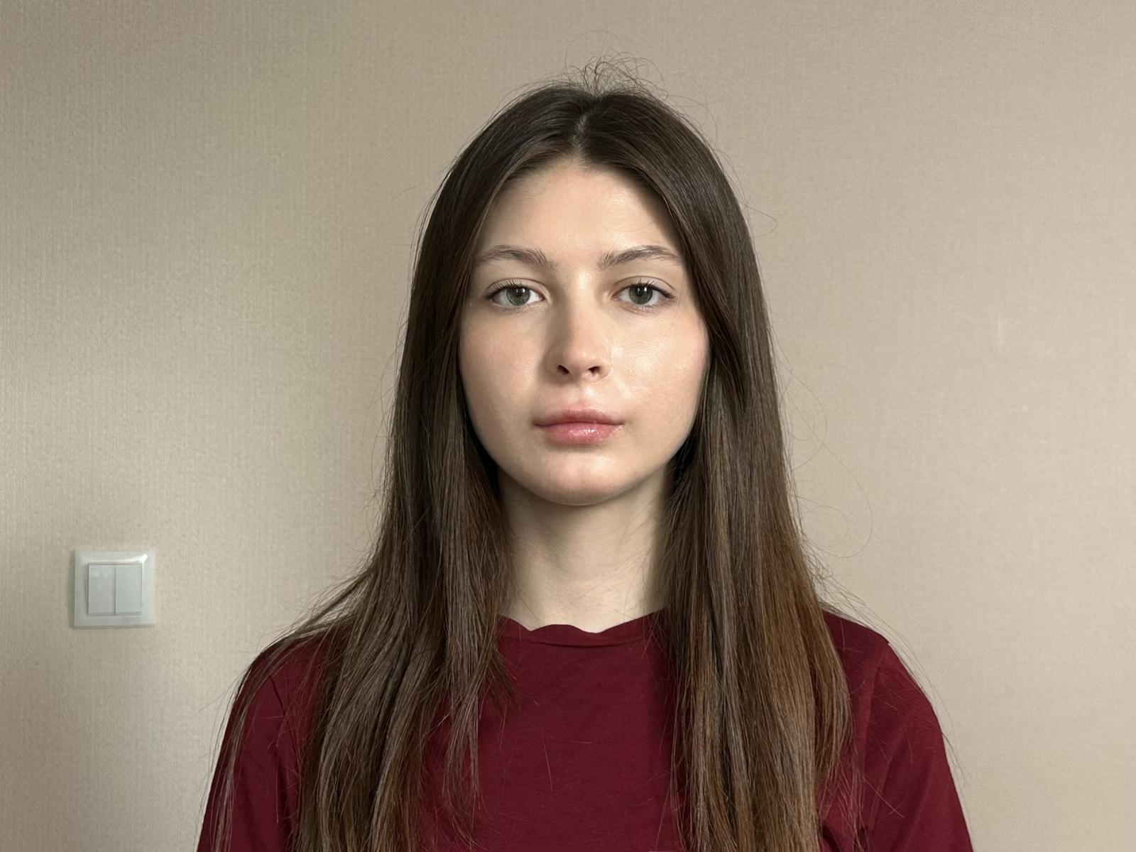 Vera Banegas, mit langen braunen Haaren, heller Haut und ohne Make-up, trägt ein rotes Hemd und hat einen neutralen Gesichtsausdruck. Auf der linken Seite befindet sich ein kleiner, weißer Lichtschalter..