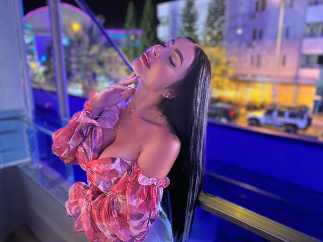 Semiramida, attraente con lunghi capelli neri, una maglietta rosa a spalle scoperte che mette in risalto il décolleté, occhi chiusi, che si tocca il mento, in una scena notturna urbana illuminata da luci al neon. Luci blu e viola la illuminano..