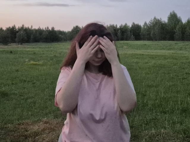 ChungWittrup, com cabelo ruivo, com as mãos cobrindo o rosto, vestindo uma camisa rosa, em pé em um campo gramado, com as mãos no rosto, expressão tímida, pôr do sol ao fundo, luz suave..
