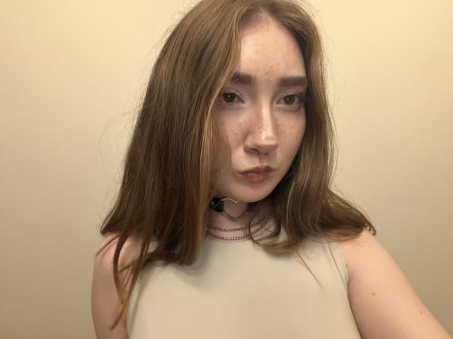 NellyAdside a la peau pâle, les cheveux bruns, un anneau au nez et des taches de rousseur. Elle porte un haut blanc sans manches avec un collier noir et regarde l'appareil photo avec une expression neutre..