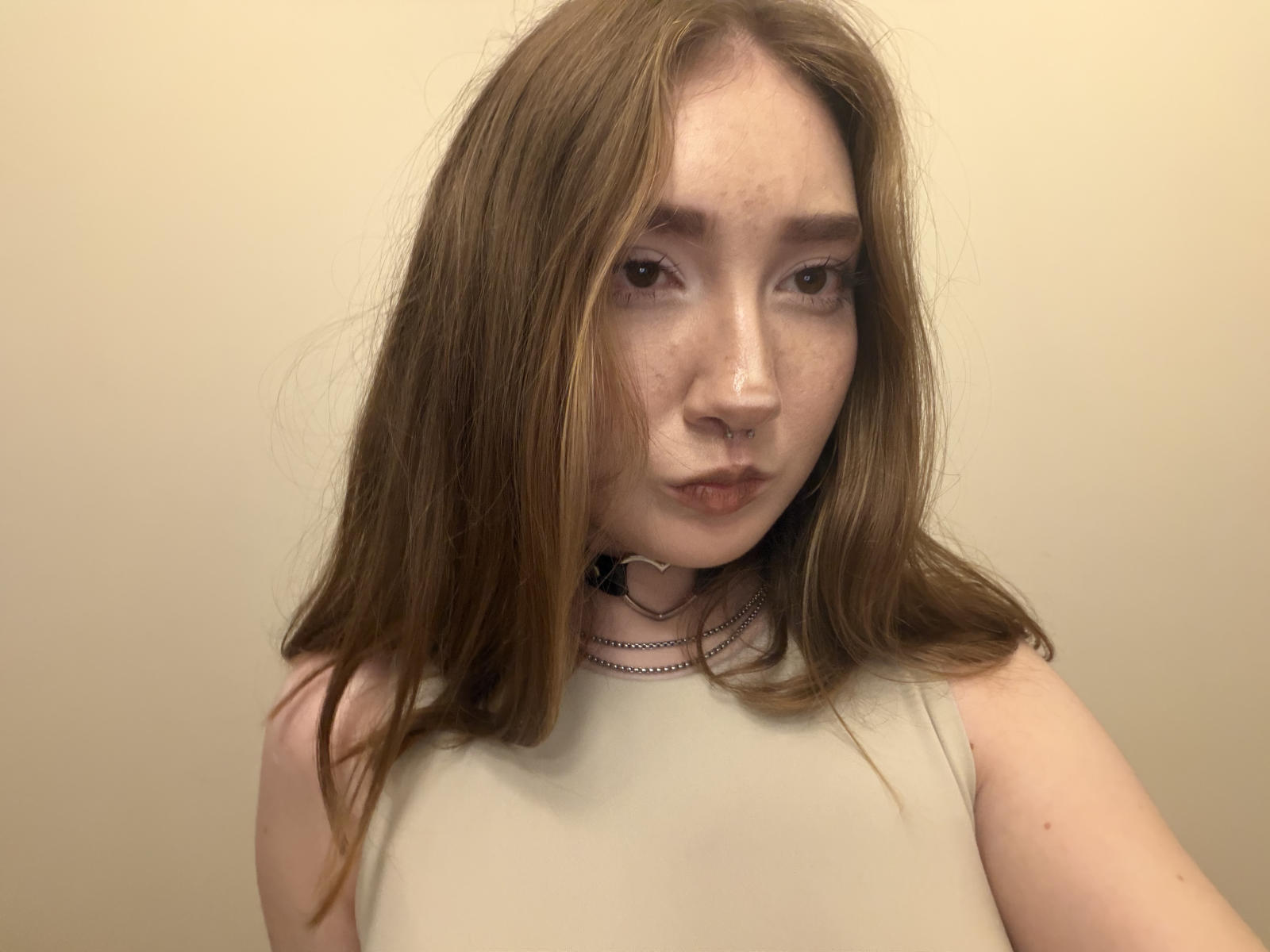 NellyAdside tem pele clara, cabelo castanho, um piercing no nariz e sardas, vestindo uma blusa branca sem mangas com um colar preto, olhando para a câmera com uma expressão neutra..