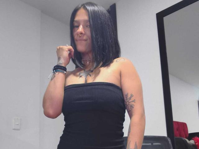 Amelie Blake, avec un haut noir sans bretelles, montrant de petites seins, un tatouage sur le bras droit, des cheveux noirs, un anneau au nez, un collier, des bracelets et un tatouage sur la poitrine, souriant légèrement..