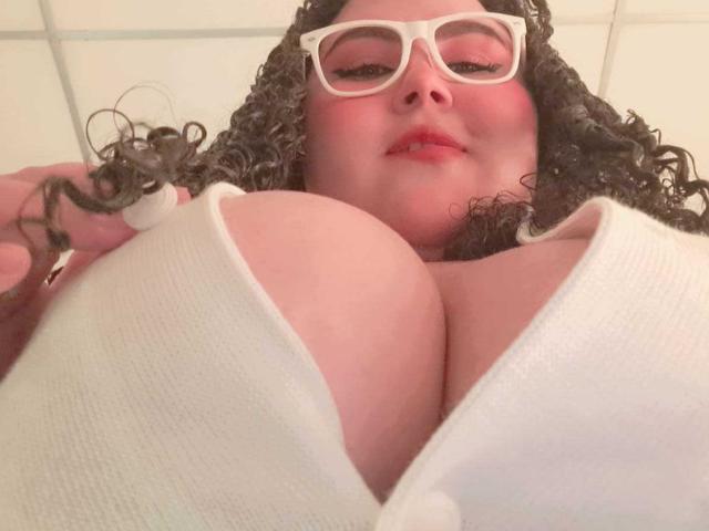 HurrenMatt with curly hair, white glasses, and red lipstick, looking down at her large, pale, bare breasts in a white, ribbed, low-cut top.