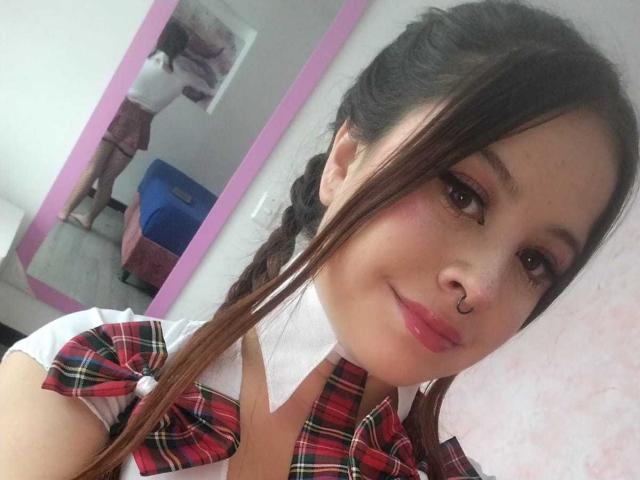 MelaniBecker has brown eyes, a nose ring, and pink lipstick. She wears a red plaid bow on a white shirt, with her braids over her shoulder. Reflection shows her back.