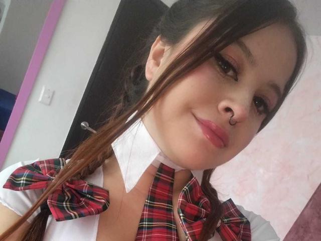 MelaniBecker with brown pigtails, wearing a red plaid schoolgirl outfit, white shirt, and matching bowtie, has pink eyeshadow, a nose ring, and glossy pink lips.