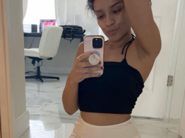 Justmxx mit kleinen Brüsten, einem schwarzen Crop-Top und rosa Hosen, macht ein Selfie vor einem Spiegel in einem weißen Raum und hält ein rosa Handy mit einem Pop-Socket..