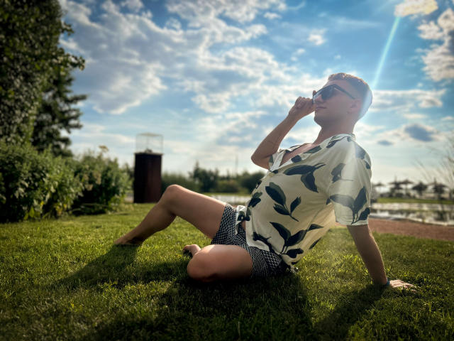 OhMyAndy, assis sur l'herbe, portant une chemise blanche avec des motifs d'oiseaux bleus, un short à carreaux noirs, des lunettes de soleil, et tenant une cigarette, avec un ciel ensoleillé et nuageux en arrière-plan..