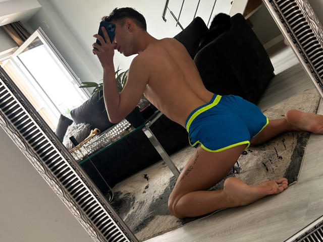 OhMyAndy kniet, ist oben ohne, trägt blaue und gelbe Shorts, hat ein Tattoo an der rechten Oberschenkel, macht ein Selfie im Spiegel, hält ein Handy, spiegelt sich in einem großen, verzierten Spiegel wider, Zimmer mit schwarzem Sofa..