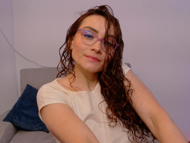 Drikka with wet, curly brown hair, purple glasses, and a white tee, smirking slightly, hands near head. Soft light on pale skin, blue pillow in gray chair.