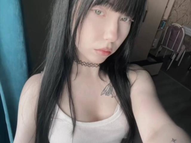 EllenChristoph, com cabelos pretos longos, pele clara, olhos verdes, top branco, colar preto, tatuagem no ombro direito, seios pequenos, sentada perto de uma cortina azul, olhando para a câmera..
