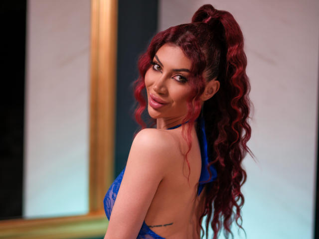 RubyMistique, con cabello rojo ondulado, lleva un sujetador de encaje azul y mira por encima del hombro con una sonrisa juguetona, mostrando su espalda y un pequeño tatuaje, en una habitación bien iluminada..
