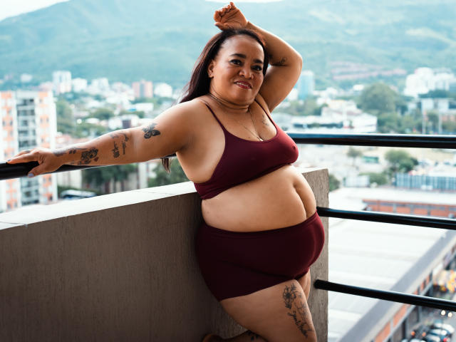 MarillinMoon è una donna latina robusta con capelli scuri, che indossa un reggiseno sportivo e dei pantaloncini color borgogna, appoggiata a un balcone, mostrando tatuaggi sul braccio e sulla coscia, e sorride..