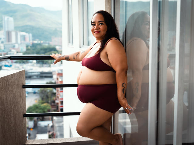 MarillinMoon, una donna formosa con capelli scuri, è in piedi su un balcone, indossa un reggiseno sportivo e pantaloncini color bordeaux, mostrando il seno e i glutei, ha tatuaggi sulle braccia ed è sorridente..