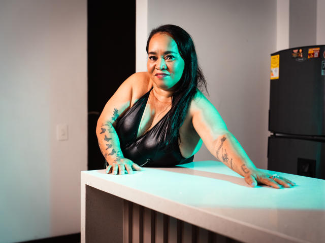 MarillinMoon with tattoos on arms, wearing a black leather halter, leaning on a white kitchen counter, looking at camera, green light on left side, black fridge on right.