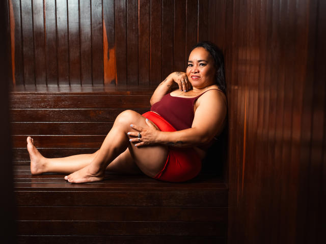 MarillinMoon, a curvy woman with medium brown skin and dark hair, sits on wooden stairs in a red tank top and red shorts, smiling with one hand on her knee.