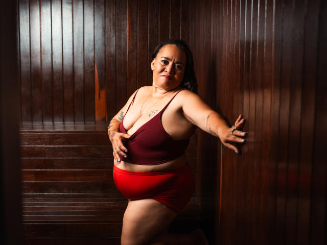 MarillinMoon with medium tits in a red bra and red panties, leaning against dark wooden walls, right hand on wall, left hand on bra, confident smile, dark hair.