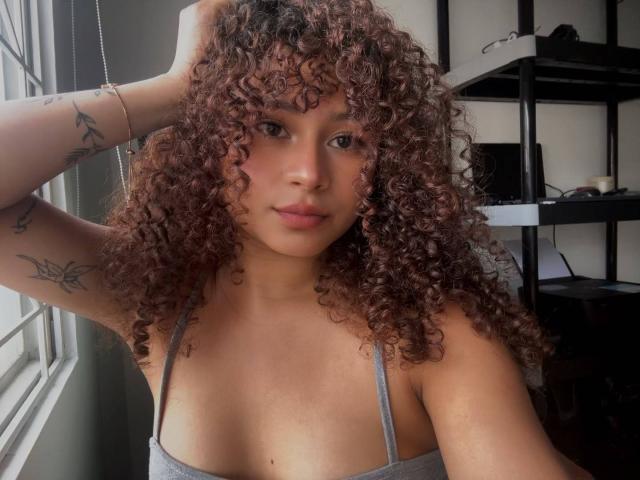 SofiaPassionne tiene el pelo castaño rizado, piel morena, pechos pequeños, tatuajes en los brazos, una camiseta gris sin mangas, una mano en el pelo y está mirando a la cámara con una ligera sonrisa..