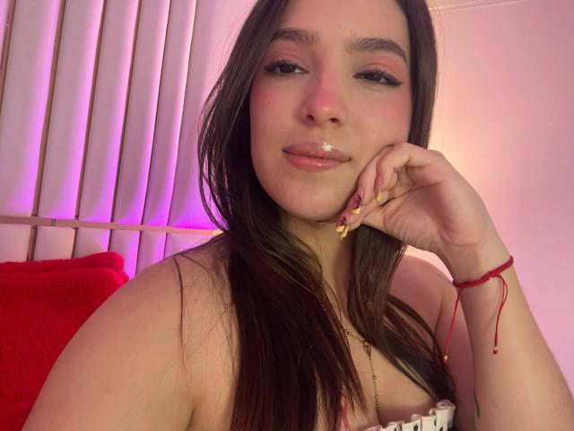 Katarina Novak, avec de longs cheveux bruns, du fard à paupières rose, des lèvres brillantes, le menton appuyé sur la main, une manucure dorée, un bracelet rouge, les épaules dénudées, un oreiller rouge, une lumière rose et violette..