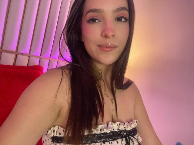 Katarina Novak, avec de longs cheveux bruns, du fard à paupières rose, des lèvres brillantes, portant un haut noir et blanc à motifs sans bretelles, avec une petite poitrine, une peau claire et un collier en or, souriant..