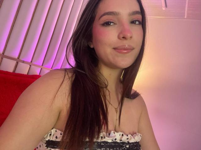 Katarina Novak, avec de longs cheveux bruns, un maquillage discret et un petit piercing sur la lèvre, porte un haut sans bretelles à pois, et sourit doucement, montrant sa petite poitrine ronde et ses joues rosées..