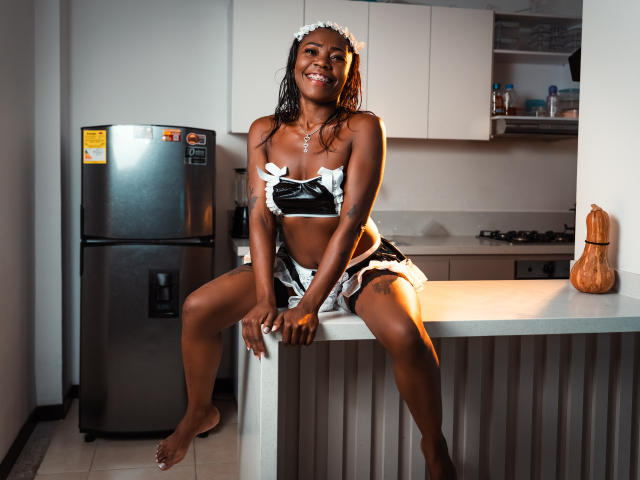 SofiiKinky, de pele escura, vestindo um uniforme de empregada francesa preto e branco, está sentada em uma bancada de cozinha, sorrindo, com os pés descalços, mostrando os seios e um rosto bonito..