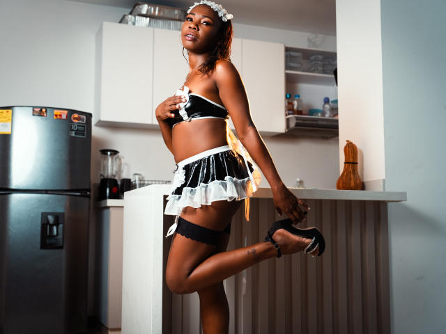 SofiiKinky in a black and white maid outfit, showing her tits, holding a yellow ribbon, with black heels and a lace garter, standing in a kitchen, one leg up.