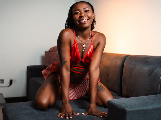 SofiiKinky, donna nera, sorridente, con un top da bikini rosso lucido, una gonna a scacchi rossi, inginocchiata su un divano grigio, con tatuaggi sulle cosce, seno di media grandezza, pelle scura, trecce corte, collana d'argento..