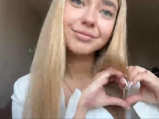 Georgine Branen, avec ses cheveux blonds, ses yeux bleus et son petit sourire, formant un cœur avec ses mains, portant un haut blanc, a l'air mignonne et détendue..