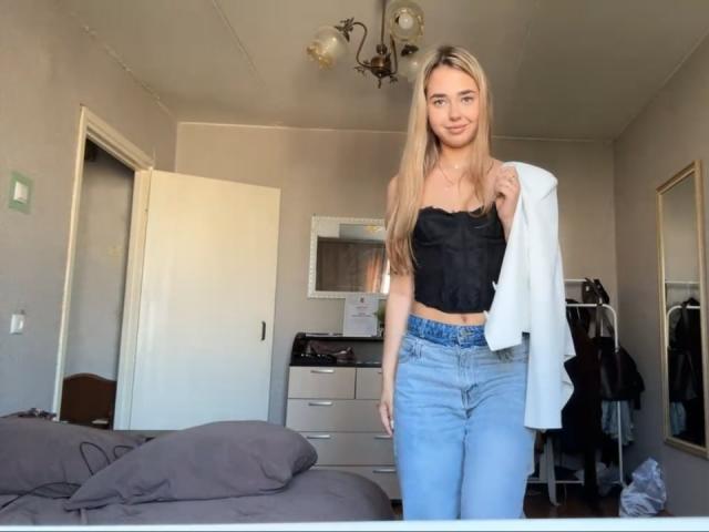 Georgine Branen, com cabelos loiros longos, seios pequenos em um top preto sem alças, segurando uma camisa branca, vestindo calças jeans azuis de cintura alta, em pé em um quarto..