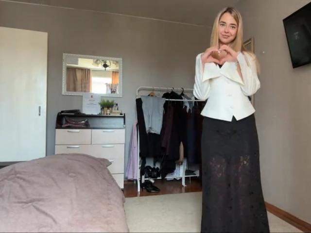 Georgine Branen, avec ses longs cheveux blonds, sa blouse blanche et sa jupe noire, se tient dans une chambre, formant un cœur avec ses mains, montrant de petites seins, et affichant un sourire mignon..