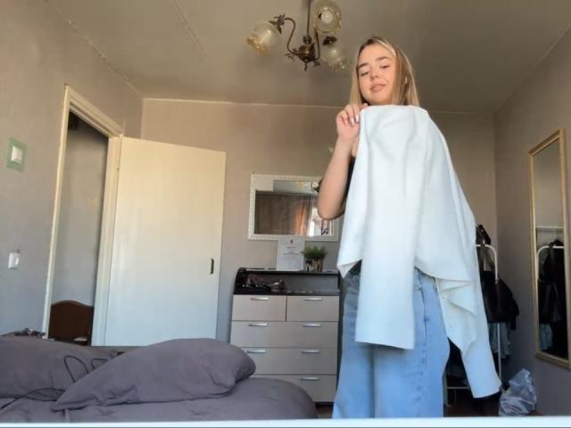 Georgine Branen, com cabelo loiro, segurando uma camisa branca, tem um traseiro pequeno, vestindo uma saia jeans azul, e está em um quarto com uma cama cinza e um armário branco..