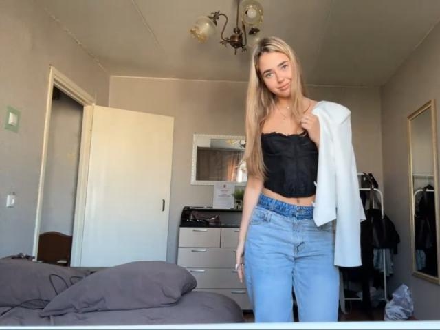 Georgine Branen, com cabelos loiros longos, um corpete preto sem alças e jeans azuis, segurando uma camisa branca, está em pé em um quarto com uma cama e um armário..