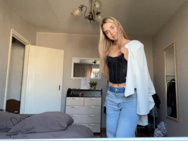 Georgine Branen, com cabelos loiros longos, seios pequenos, vestindo um top preto sem alças, jeans azuis e uma camisa branca jogada sobre o ombro, está em pé em um quarto com um lustre acima..