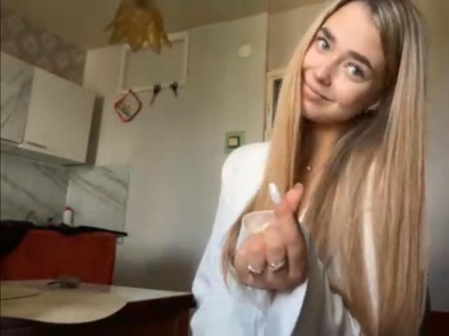 Georgine Branen mit langem, blondem Haar, weißem Hemd und erhobenem Finger, die in einer Küche ein leichtes Lächeln zeigt. Ihre Brüste sind klein, und ihre Nägel sind weiß lackiert..