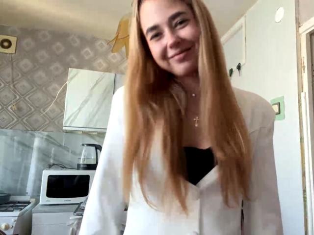 Georgine Branen, com cabelos longos, lisos e loiros, sorrindo, vestindo uma jaqueta branca sobre uma blusa preta, está em uma cozinha, mostrando seus seios levemente através da jaqueta..