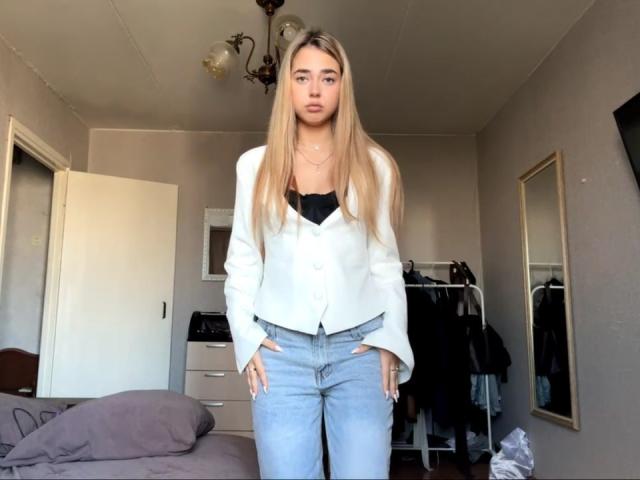 Georgine Branen, avec de longs cheveux blonds, une veste blanche, un soutien-gorge noir, un jean bleu clair, debout dans une chambre, les mains dans les poches, regardant la caméra, lumière douce, expression neutre, maquillage discret..