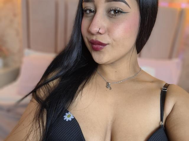 SophieeCollen, con cabello negro largo, un piercing en la nariz y lápiz labial oscuro, lleva un sujetador negro con una pequeña flor, mostrando sus grandes senos, y un collar de plata..