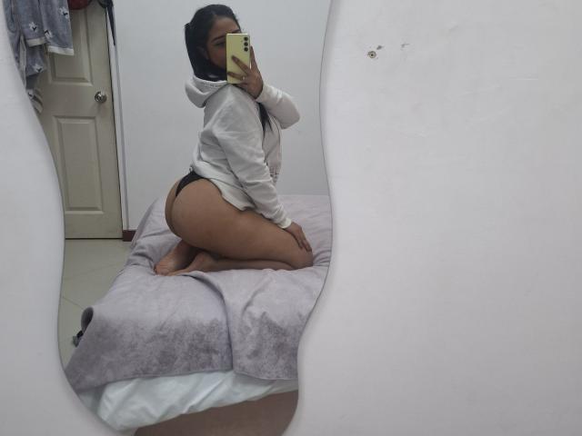 SophieeCollen knielend op een bed, gekleed in een witte hoodie en een zwarte string, waarbij haar billen zichtbaar zijn, maakt een selfie in de spiegel met een gele telefoon..