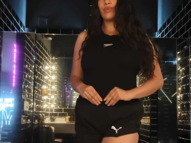 Rachel Wilson heeft lang zwart haar, draagt een zwart topje zonder mouwen en zwarte korte broek met een wit Puma-logo. Ze staat met haar handen op haar heupen, waarbij haar borsten zichtbaar zijn..