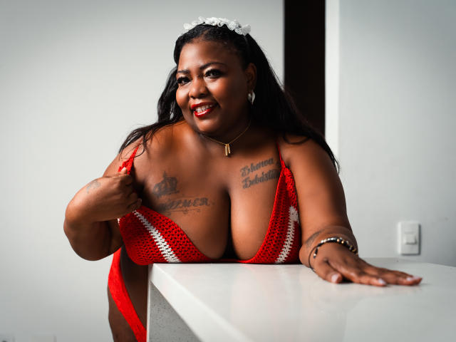 ZoeSmitth with big boobs, wearing a red knit top, pulling it down to show her tits, smiling, dark skin, tattoos on chest, white flower in hair, standing by white counter.