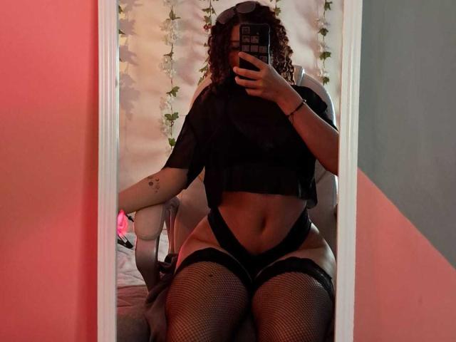 Sofia Jackson, con i capelli ricci, indossa un top corto nero, biancheria intima nera, calze a rete, tiene in mano un telefono, è un selfie allo specchio, ha un tatuaggio visibile sul braccio destro, le pareti sono rosse e grigie, è seduta su una sedia..