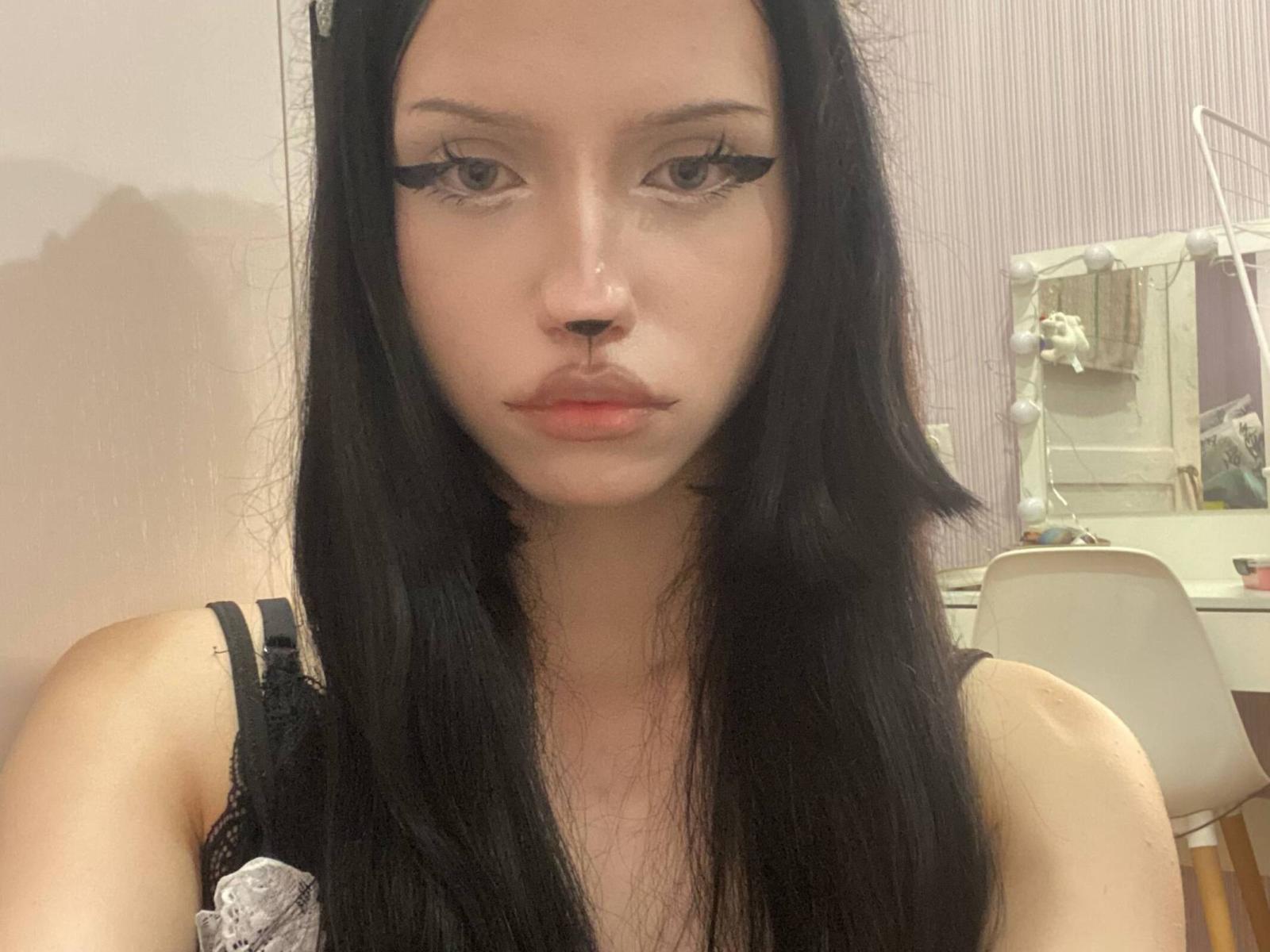 CherelleGess ha lunghi capelli neri, un trucco occhi nero molto marcato e labbra piene, e indossa una camicetta nera in pizzo. Guarda la fotocamera con un'espressione seria..