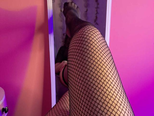 SofiaJackson's legs in black fishnet stockings, reflected in a mirror, showing her foot up, pink and orange walls, soft purple and blue lighting, in a sensual pose.