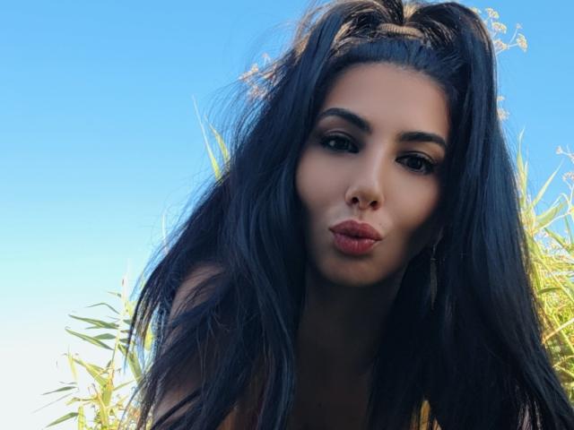 BrunetteBabe69 a de longs cheveux foncés, des lèvres pulpeuses, et elle fait une moue de baiser, avec un ciel bleu clair et des feuilles vertes en arrière-plan..