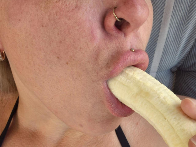 JennaStorm, attraente con l'anello al naso, gli orecchini e il piercing al naso, sta succhiando una banana con la bocca aperta, mostrando le sue labbra rosa e la pelle leggermente lentigginosa..