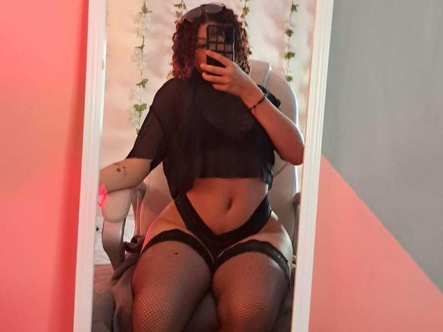 Sofia Jackson, con cabello rojo rizado, una blusa negra transparente, ropa interior negra y medias de rejilla, sentada en una silla, tomando una selfie frente al espejo, mostrando sus curvas y su abdomen suave..
